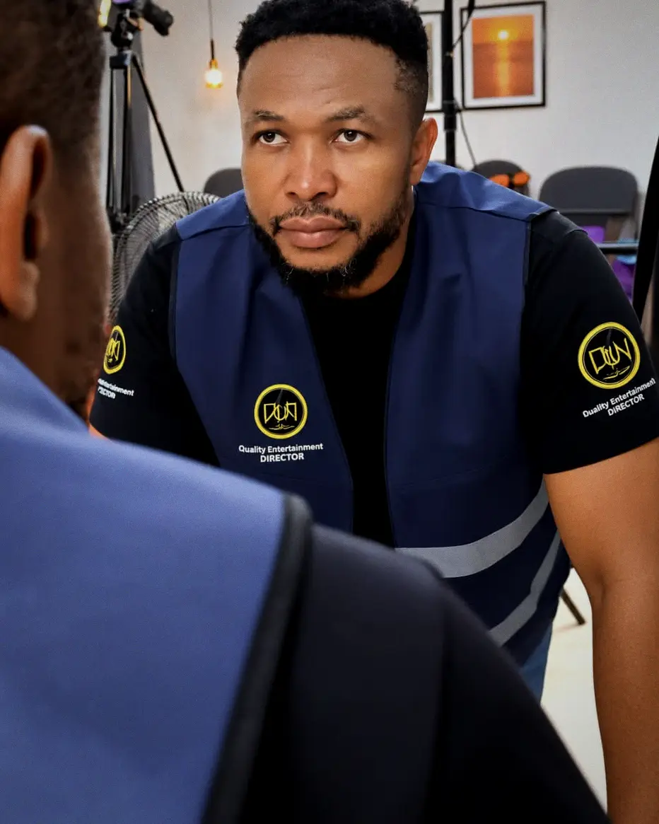 David Nnaji in a director's vest leans in to listen to a crew member on set, lights and camera soft behind him.
