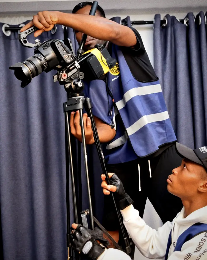 A senior crew member mounts a camera onto a tripod while a younger crew member in grip gloves steadies the legs and watches.